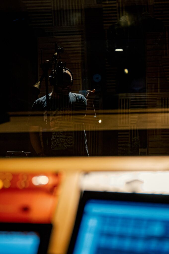 Through glass of musician in headphones recording song into microphone in modern music studio